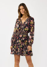 [Color: Black/Rust] A front facing image of a brunette model wearing a black floral mini dress with a smocked waist, v-neckline, and long sleeves with smocked cuffs. A feminine and flirty mini dress for weddings, holidays, or other special occasions. 