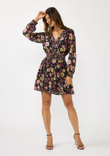 [Color: Black/Rust] A front facing image of a brunette model wearing a black floral mini dress with a smocked waist, v-neckline, and long sleeves with smocked cuffs. A feminine and flirty mini dress for weddings, holidays, or other special occasions. 