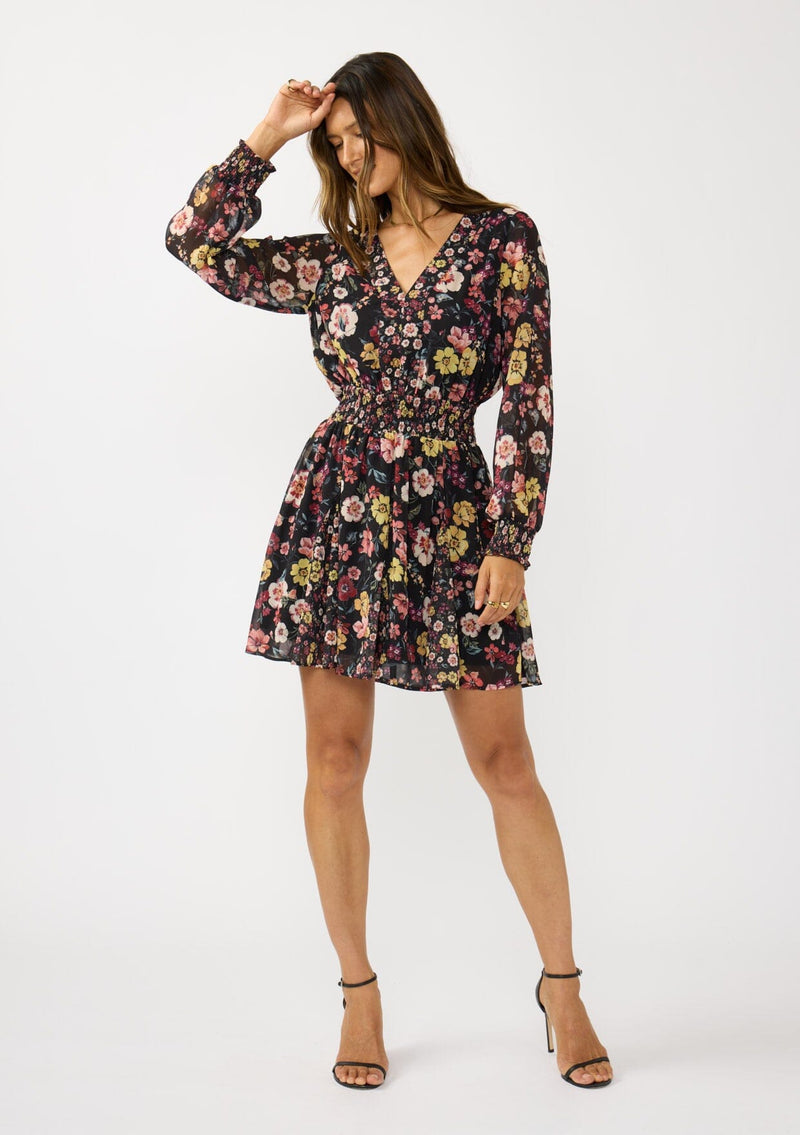 [Color: Black/Rust] A front facing image of a brunette model wearing a black floral mini dress with a smocked waist, v-neckline, and long sleeves with smocked cuffs. A feminine and flirty mini dress for weddings, holidays, or other special occasions. 