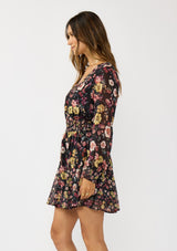 [Color: Black/Rust] A side facing image of a brunette model wearing a black floral mini dress with a smocked waist, v-neckline, and long sleeves with smocked cuffs. A feminine and flirty mini dress for weddings, holidays, or other special occasions. 