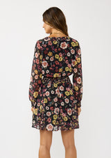 [Color: Black/Rust] A back facing image of a brunette model wearing a black floral mini dress with a smocked waist, v-neckline, and long sleeves with smocked cuffs. A feminine and flirty mini dress for weddings, holidays, or other special occasions. 