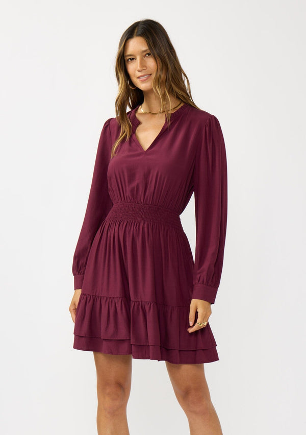 [Color: Merlot] A front facing image of a brunette model wearing a burgundy long sleeve mini dress with a split v-neckline, smocked waist, and a layered ruffle hem. Effortlessly chic and elegant for special occasions. 