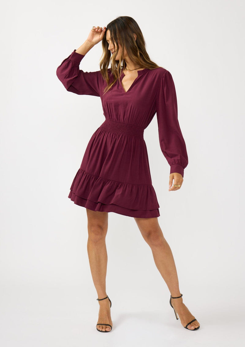 [Color: Merlot] A front facing image of a brunette model wearing a burgundy long sleeve mini dress with a split v-neckline, smocked waist, and a layered ruffle hem. Effortlessly chic and elegant for special occasions. 