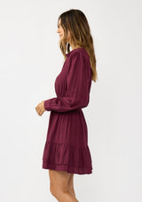 [Color: Merlot] A  side facing image of a brunette model wearing a burgundy long sleeve mini dress with a split v-neckline, smocked waist, and a layered ruffle hem. Effortlessly chic and elegant for special occasions. 