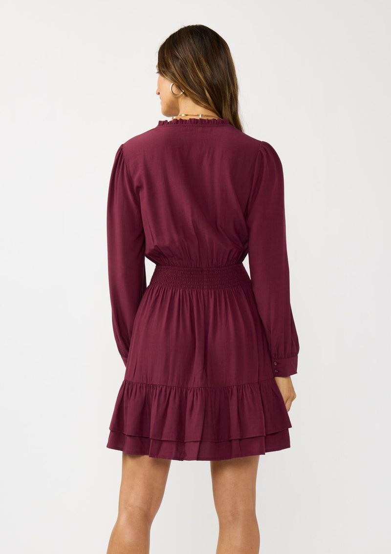 [Color: Merlot] A back facing image of a brunette model wearing a burgundy long sleeve mini dress with a split v-neckline, smocked waist, and a layered ruffle hem. Effortlessly chic and elegant for special occasions. 