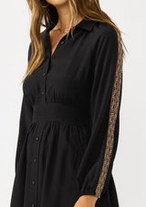 [Color: Black] A side facing image of a brunette model wearing a black dress with embroidered sleeves, a button front, a collared v-neckline, and side pockets. Effortlessly boho and casual for day to night. 