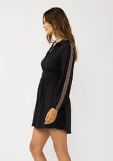 [Color: Black] A side facing image of a brunette model wearing a black dress with embroidered sleeves, a button front, a collared v-neckline, and side pockets. Effortlessly boho and casual for day to night. 