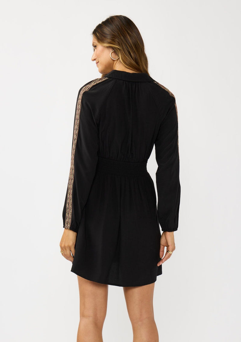 [Color: Black] A back facing image of a brunette model wearing a black dress with embroidered sleeves, a button front, a collared v-neckline, and side pockets. Effortlessly boho and casual for day to night. 