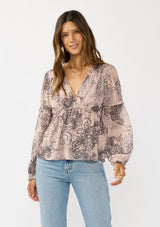 [Color: Natural/Brown] A front facing image of a brunette model wearing a nude brown floral blouse with a deep v-neckline, and ruffle accents on the front and sleeves. A classic boho top for casual outings and special occasions.  