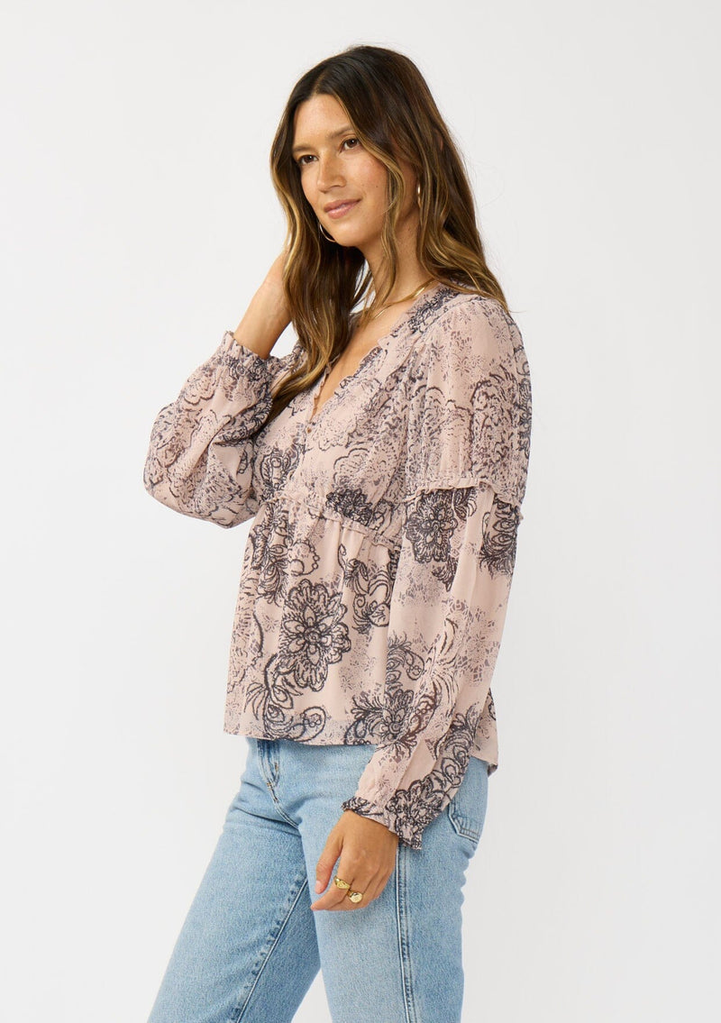 [Color: Natural/Brown] A side facing image of a brunette model wearing a nude brown floral blouse with a deep v-neckline, and ruffle accents on the front and sleeves. A classic boho top for casual outings and special occasions.  