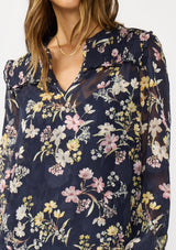 [Color: Navy/Yellow] A detail image of a brunette model wearing a navy floral burnout blouse with smocked yoke, split v-neckline, and ruffle trims. 