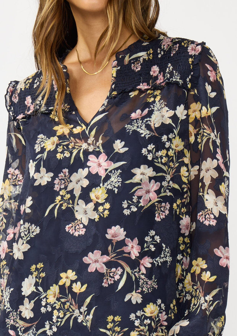 [Color: Navy/Yellow] A detail image of a brunette model wearing a navy floral burnout blouse with smocked yoke, split v-neckline, and ruffle trims. 