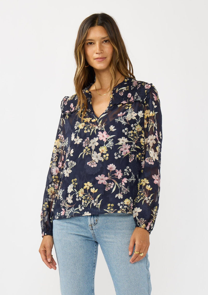 [Color: Navy/Yellow] A front facing image of a brunette model wearing a navy floral burnout blouse with smocked yoke, split v-neckline, and ruffle trims. 