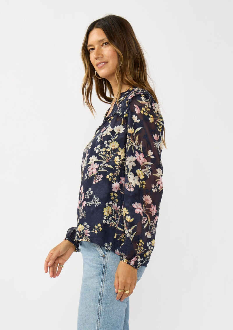 [Color: Navy/Yellow] A side facing image of a brunette model wearing a navy floral burnout blouse with smocked yoke, split v-neckline, and ruffle trims. 
