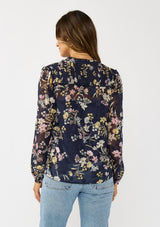 [Color: Navy/Yellow] A back facing image of a brunette model wearing a navy floral burnout blouse with smocked yoke, split v-neckline, and ruffle trims. 