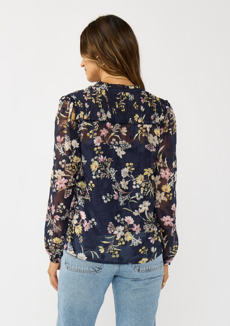 [Color: Navy/Yellow] A back facing image of a brunette model wearing a navy floral burnout blouse with smocked yoke, split v-neckline, and ruffle trims. 