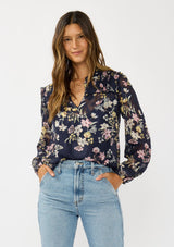 [Color: Navy/Yellow] A front facing image of a brunette model wearing a navy floral burnout blouse with smocked yoke, split v-neckline, and ruffle trims. 