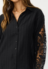 [Color: Black] A detail image of a blonde model wearing a striped jacquard black shirt with lace cutouts, a button front, and collared neckline. A cool and polished blouse for the fall season. 
