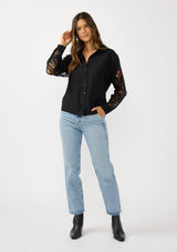 [Color: Black] A front facing image of a blonde model wearing a striped jacquard black shirt with lace cutouts, a button front, and collared neckline. A cool and polished blouse for the fall season. 