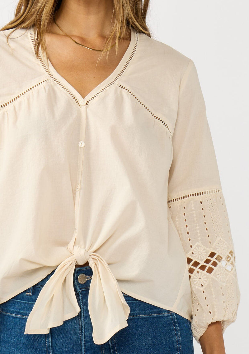 [Color: Natural] A detail  image of a blonde model wearing an ivory lightweight cotton-blend top with a tie front detail, button front, and 3/4 length sleeves with embroidered and cutout details. 