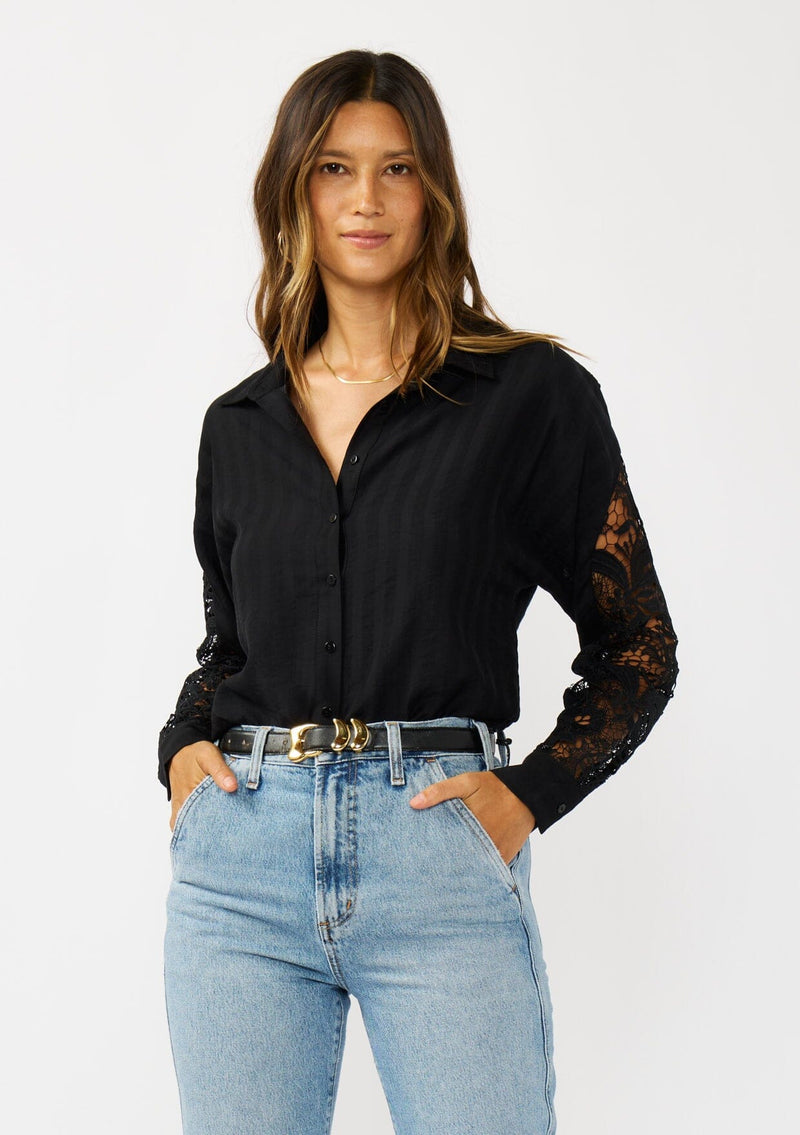 [Color: Black] A front facing image of a blonde model wearing a striped jacquard black shirt with lace cutouts, a button front, and collared neckline. A cool and polished blouse for the fall season. 