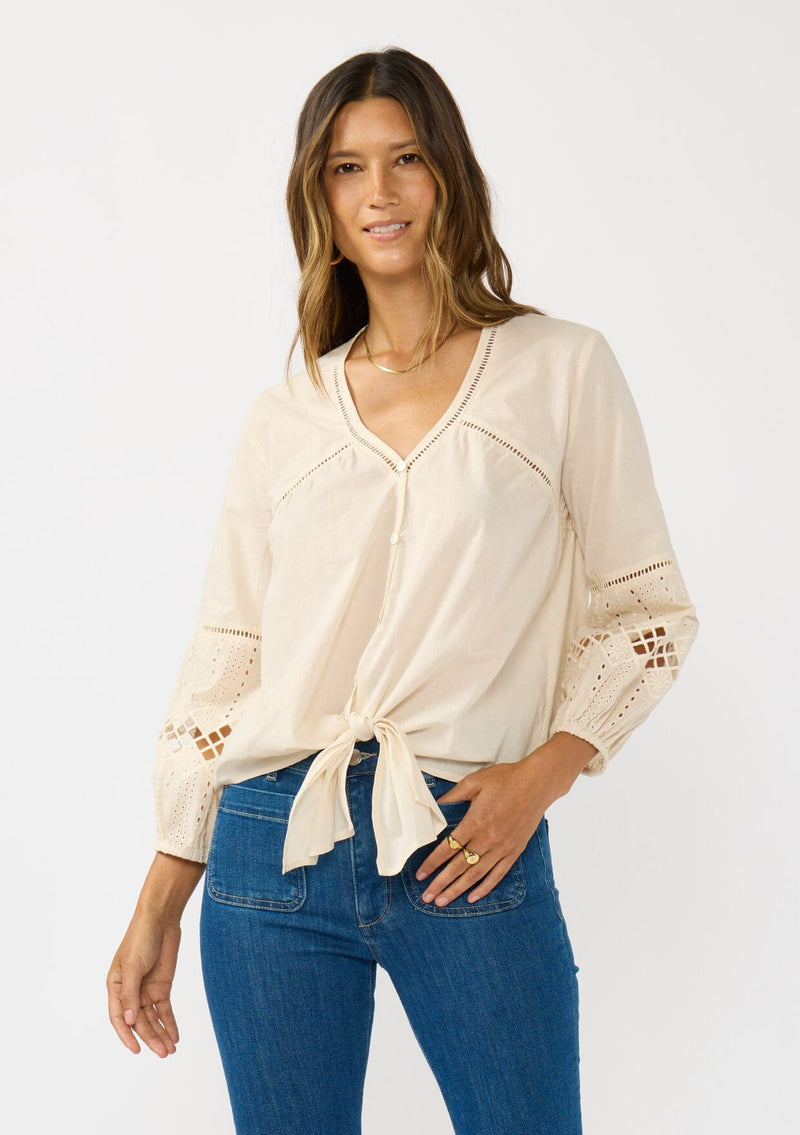 [Color: Natural] A front facing image of a blonde model wearing an ivory lightweight cotton-blend top with a tie front detail, button front, and 3/4 length sleeves with embroidered and cutout details. 