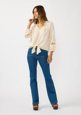 [Color: Natural] A front facing image of a blonde model wearing an ivory lightweight cotton-blend top with a tie front detail, button front, and 3/4 length sleeves with embroidered and cutout details. 