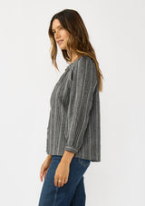 [Color: Charcoal] A side facing image of a brunette model wearing a dark grey striped blouse with long sleeves, a split v-neckline, and button front. 