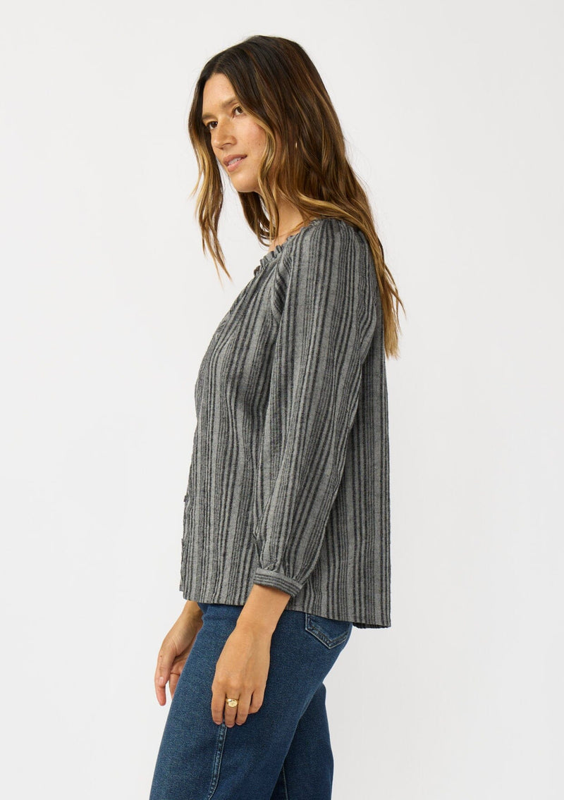 [Color: Charcoal] A side facing image of a brunette model wearing a dark grey striped blouse with long sleeves, a split v-neckline, and button front. 