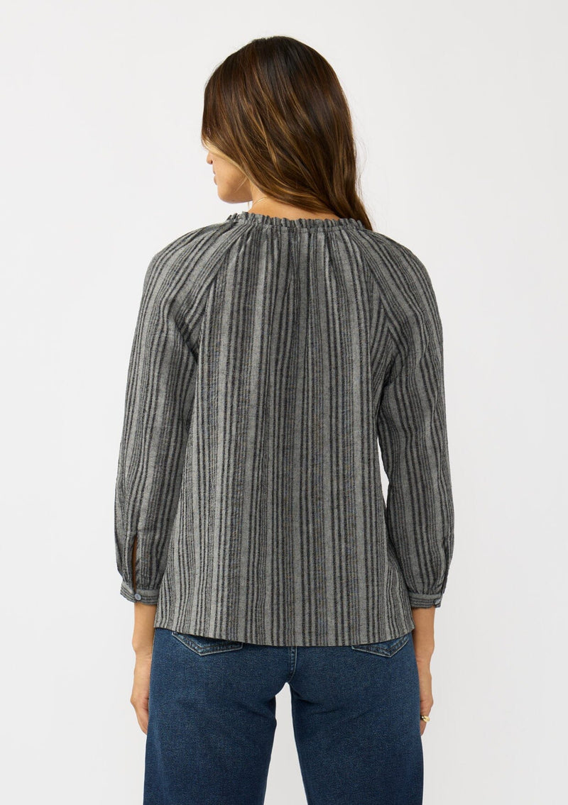 [Color: Charcoal] A back facing image of a brunette model wearing a dark grey striped blouse with long sleeves, a split v-neckline, and button front. 