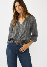 [Color: Charcoal] A front facing image of a brunette model wearing a dark grey striped blouse with long sleeves, a split v-neckline, and button front. 