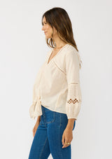 [Color: Natural] A side facing image of a blonde model wearing an ivory lightweight cotton-blend top with a tie front detail, button front, and 3/4 length sleeves with embroidered and cutout details. 