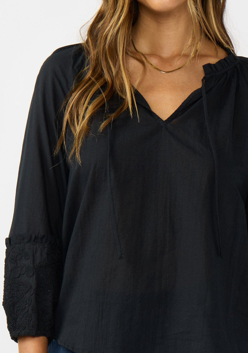 [Color: Black] A detail image of a brunette model wearing a black cotton blouse with tonal embroidered 3/4 sleeves and a split v-neckline with tie detail. 