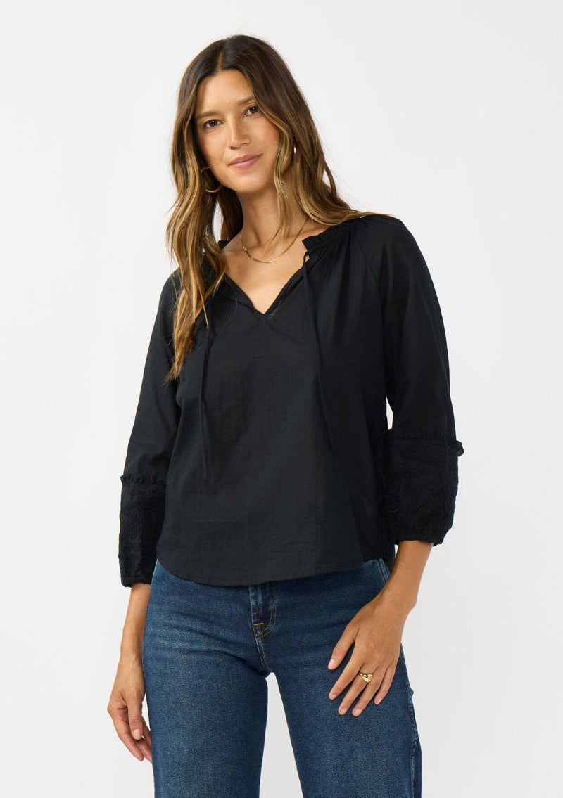 [Color: Black] A front facing image of a brunette model wearing a black cotton blouse with tonal embroidered 3/4 sleeves and a split v-neckline with tie detail. 