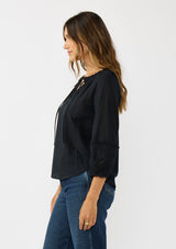 [Color: Black] A side facing image of a brunette model wearing a black cotton blouse with tonal embroidered 3/4 sleeves and a split v-neckline with tie detail. 