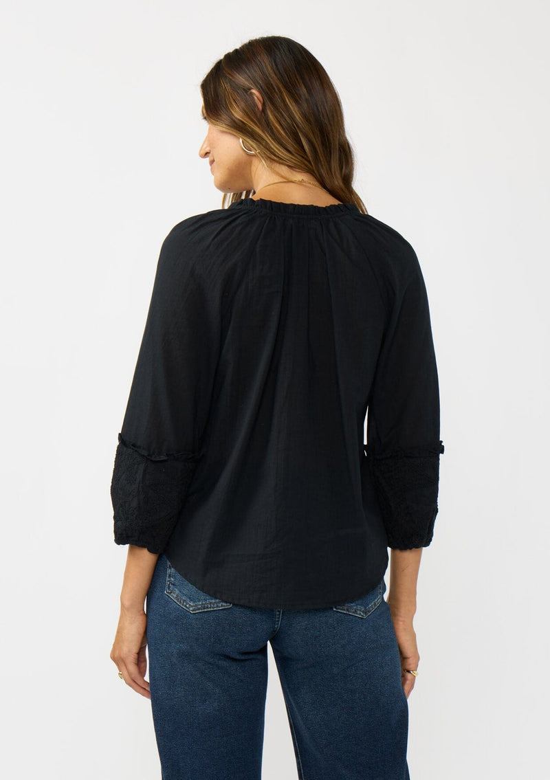 [Color: Black] A back facing image of a brunette model wearing a black cotton blouse with tonal embroidered 3/4 sleeves and a split v-neckline with tie detail. 