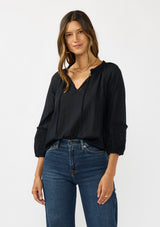 [Color: Black] A front facing image of a brunette model wearing a black cotton blouse with tonal embroidered 3/4 sleeves and a split v-neckline with tie detail. 