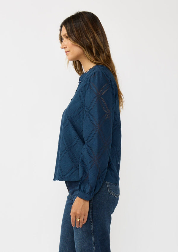 [Color: Navy] A side facing image of a brunette model wearing a navy blue button up blouse with a diamond lattice pattern and long sleeves with button cuffs. A versatile top for the office and weekend casual outings. 