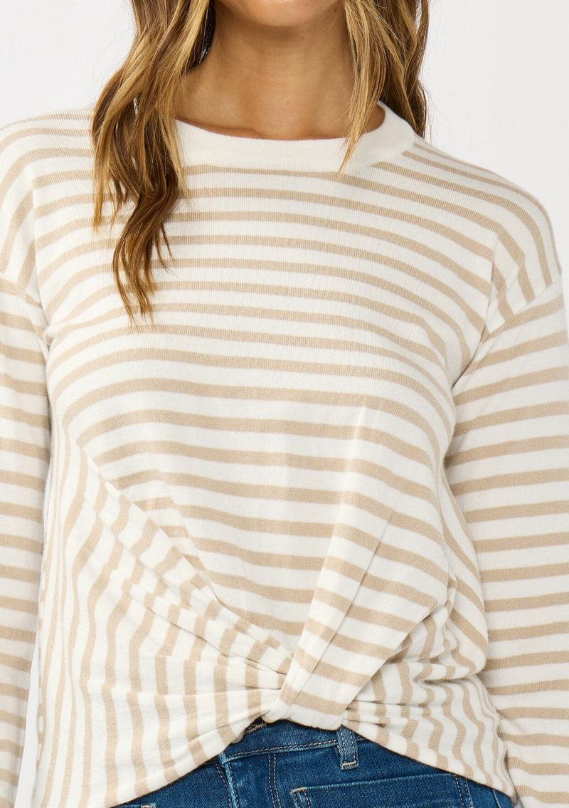 [Color: Ivory/Tan] A detail image of a brunette model wearing a classic ivory and tan brown striped knit pullover sweater. With long sleeves, a drop shoulder, a crew neckline, and a twist front detail at the waist. 