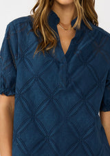 [Color: Navy] A detail image of a brunette model wearing a navy puff sleeve top with a diamond lattice pattern, ruffled details, and a split v-neckline. 