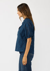 [Color: Navy] A side facing image of a brunette model wearing a navy puff sleeve top with a diamond lattice pattern, ruffled details, and a split v-neckline. 