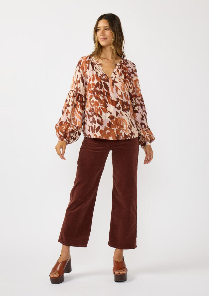 [Color: Nude/Brown] A front facing image of a brunette model wearing a nude and brown abstract print blouse with long tiered sleeves and a split v-neckline with button detail. A standout top for the fall season. 