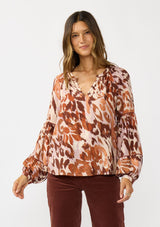 [Color: Nude/Brown] A front facing image of a brunette model wearing a nude and brown abstract print blouse with long tiered sleeves and a split v-neckline with button detail. A standout top for the fall season. 
