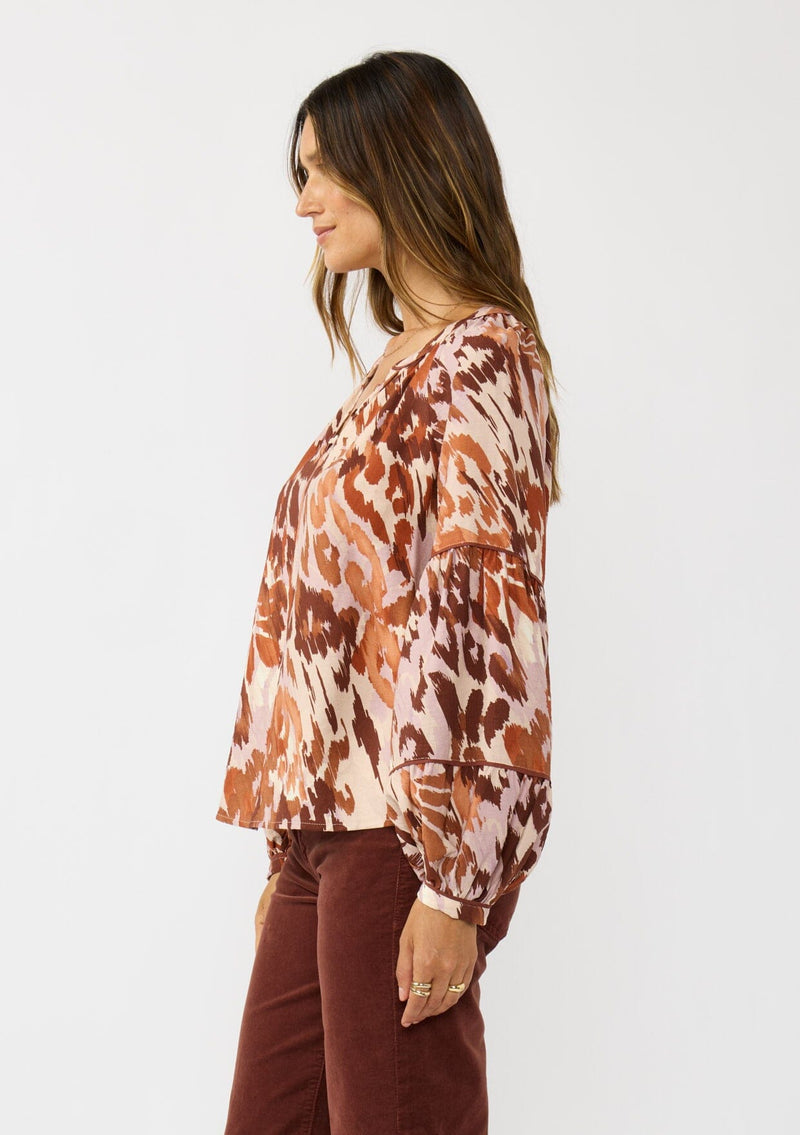 [Color: Nude/Brown] A  side facing image of a brunette model wearing a nude and brown abstract print blouse with long tiered sleeves and a split v-neckline with button detail. A standout top for the fall season. 
