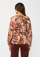 [Color: Nude/Brown] A back facing image of a brunette model wearing a nude and brown abstract print blouse with long tiered sleeves and a split v-neckline with button detail. A standout top for the fall season. 
