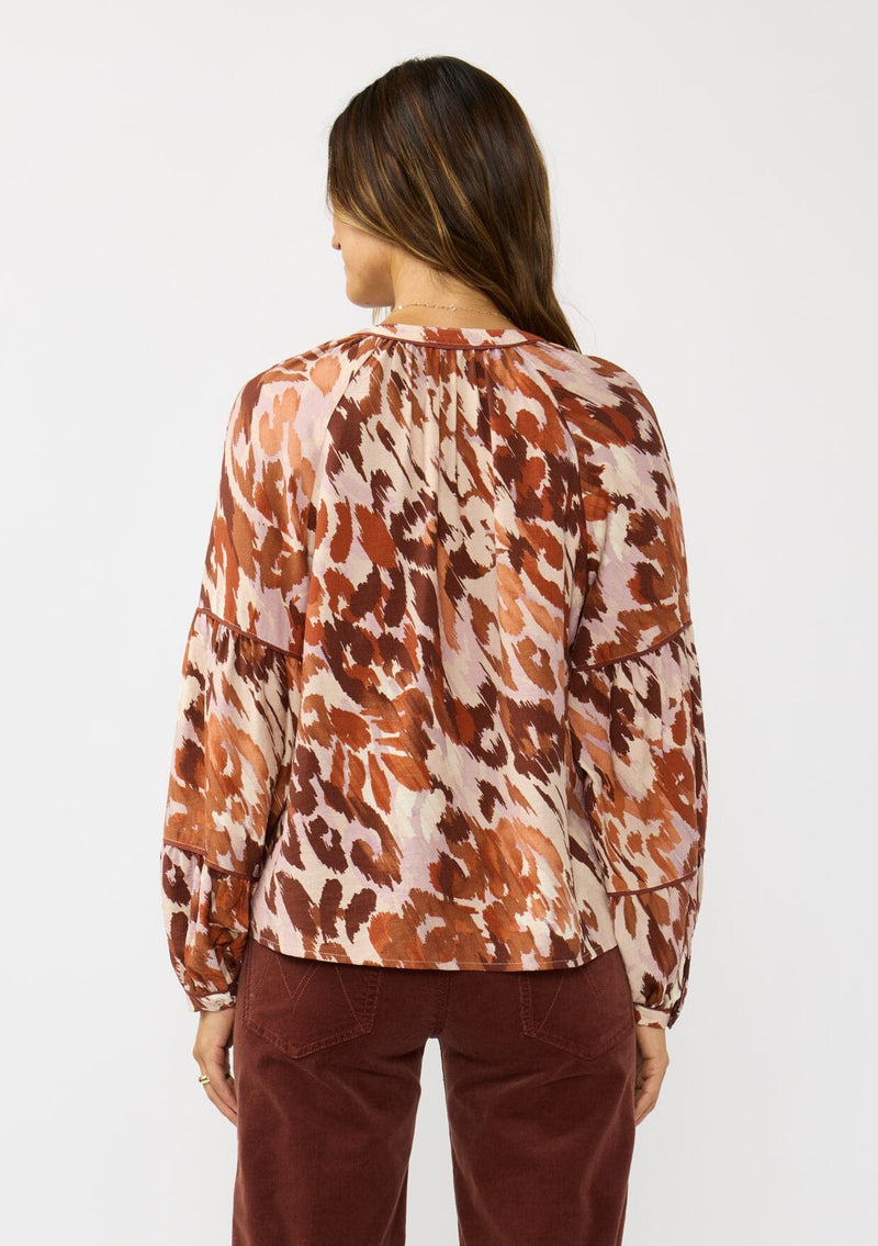 [Color: Nude/Brown] A back facing image of a brunette model wearing a nude and brown abstract print blouse with long tiered sleeves and a split v-neckline with button detail. A standout top for the fall season. 