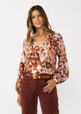 [Color: Nude/Brown] A front facing image of a brunette model wearing a nude and brown abstract print blouse with long tiered sleeves and a split v-neckline with button detail. A standout top for the fall season. 