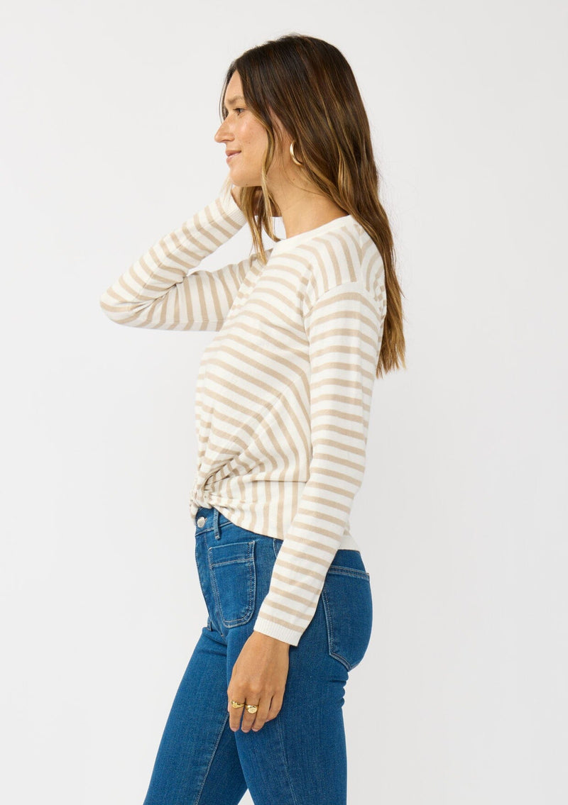 [Color: Ivory/Tan] A side facing image of a brunette model wearing a classic ivory and tan brown striped knit pullover sweater. With long sleeves, a drop shoulder, a crew neckline, and a twist front detail at the waist. 