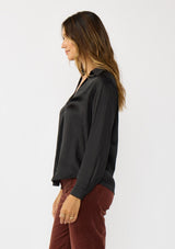 [Color: Black] A side facing image of a brunette model wearing a silky poly satin blouse with a split v-neckline, collar, and long sleeves with button cuffs. 