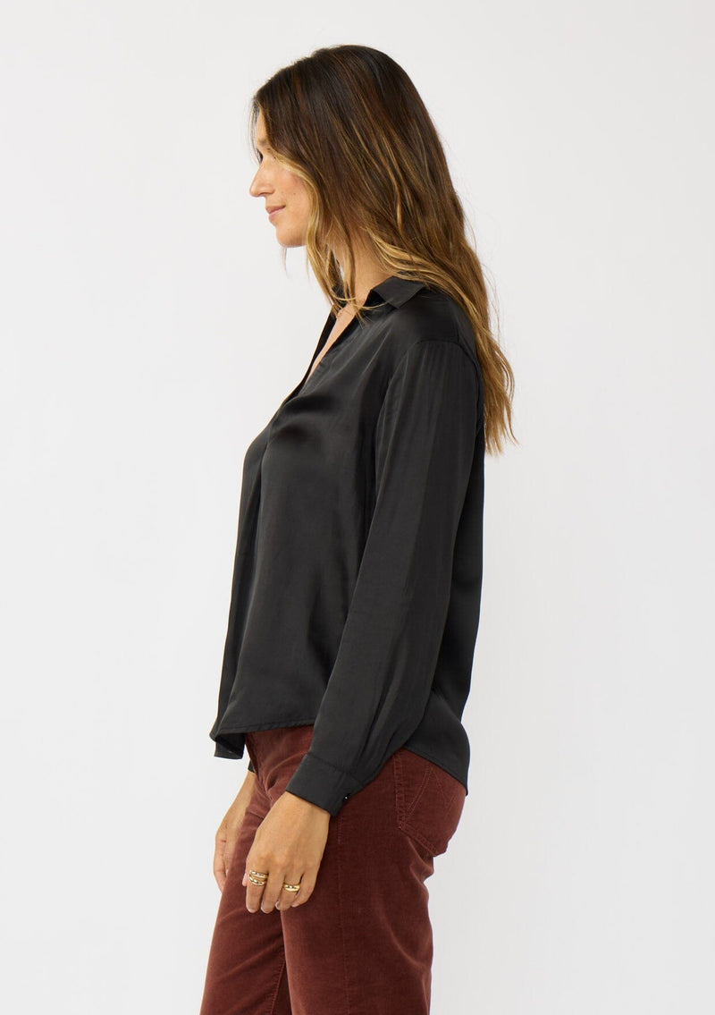 [Color: Black] A side facing image of a brunette model wearing a silky poly satin blouse with a split v-neckline, collar, and long sleeves with button cuffs. 